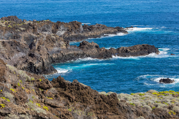 Formazione rocciosa vulcanica, scogliere di lava nera sulla riva rocciosa con onde bianche che si infrangono sull'Oceano Atlantico. Sfondo cielo blu. La Palma, Isole Canarie
. - Foto, immagini