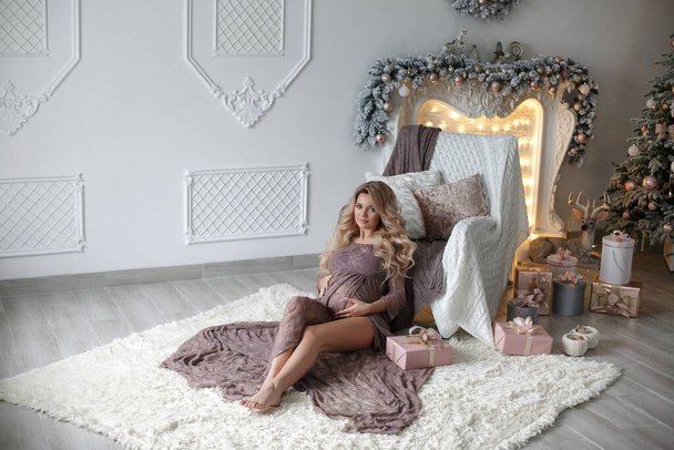 Young beautiful pregnant woman smiling and happy looking camera, hair styling makeup, white silver color on background of Christmas tree and fireplace bokeh.Portrait of beautiful pregnant woman next to christmas tree.Expectation and holiday concept. - Photo, image