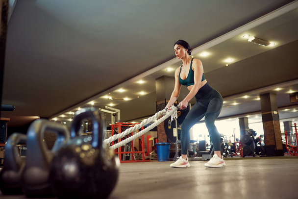 Caucasian fit woman dressed in sportsoutfit preparing for workout with battle ropes at gym - Foto, afbeelding