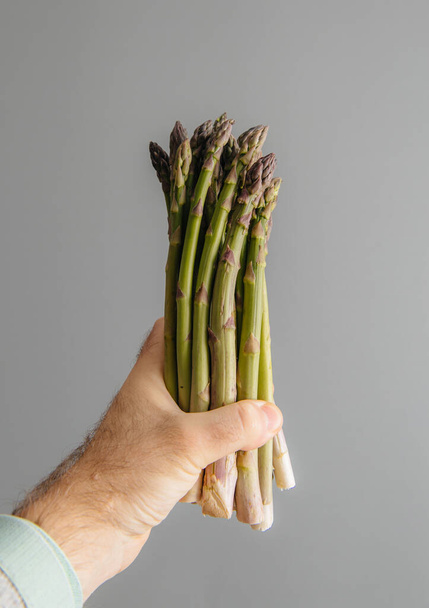 Male hand holding against gray background fresh bio organic asparagus - Foto, afbeelding