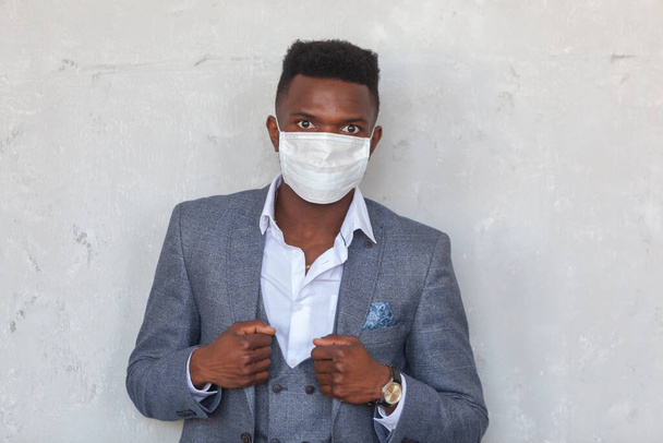 COVID-19. Confident young African man is wearing a protective medical face mask to prevent infection with coronavirus and pandemic standing in a grey suit against a concrete wall - Photo, Image