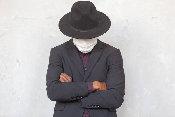 COVID-19. African man with his face hidden under black hat is wearing a protective medical face mask to prevent infection with coronavirus and pandemic standing with crossed arms in a suit against a - Photo, image