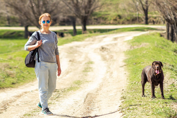 Γυναίκα με γυαλιά ηλίου που μένει στο δρόμο, με σοκολάτα σκύλου Labrador retriever. - Φωτογραφία, εικόνα