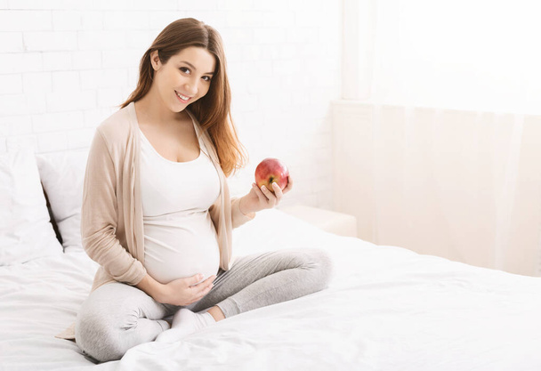 Jonge zwangere vrouw met rode appel in bed - Foto, afbeelding