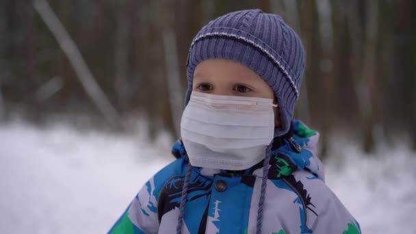 A child (boy) in winter overalls and a hat stands in the winter forest with a medical mask on his face, looks at the camera and removes the mask from his face, the end of the epidemic and self-isolati - Metraje, vídeo