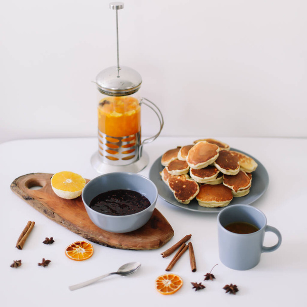 Lush pancakes and teapot with spices, cup of tea with orange, cinnamon, anise. Tasty breakfast on white background. Good morning - Photo, image