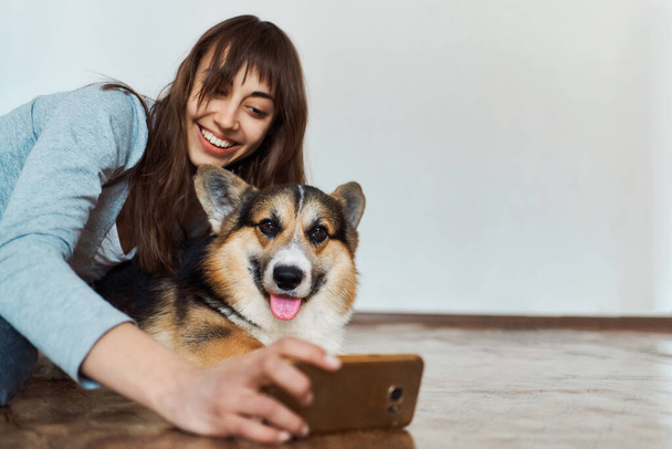 alegre sorrindo jovem mulher abraçando bonito galês cão Corgi e tirar selfie com animal de estimação na câmera do smartphone. Conceito ficar em casa, amizade com o cão, tirar fotos
. - Foto, Imagem