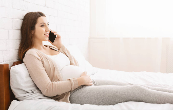 Cute pregnant woman talking on phone, lying in bed at home - Fotó, kép