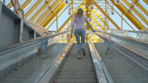 A young woman climbing up the moving escalator. Rear view, follow shot. Blond woman with long hair inside modern building - Foto, Bild