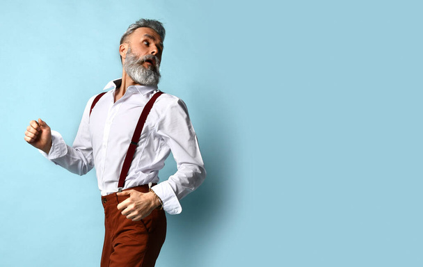 Gray-haired male in white shirt, brown pants, suspenders, black loafers. He runs looking back, posing sideways on blue background - Photo, Image