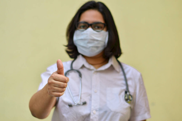 Портрет молодої медичної допомоги працівниці з великими пальцями вгору з стетоскопом на плечах одягнені в хірургічну маску, щоб захистити себе від пандемії Корони (COVID-19) на жовтому тлі. - Фото, зображення