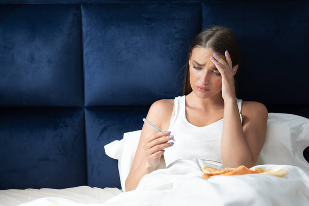Mujer enferma con fiebre que mide la temperatura sentada en la cama
 - Foto, Imagen