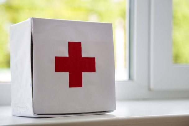 White first aid kit with red cross indoors on windowsill on blurred background. Healthcare concept. - Photo, Image