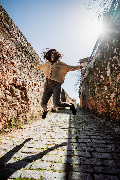 Young woman is jumping with happiness between historical walls on a narrow alleyway. Tourism and Travel concept. - Photo, Image