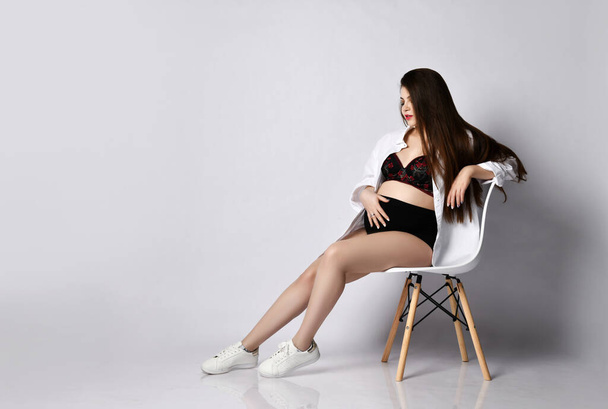 Young beautiful slim pregnant woman with long hair in white shirt sitting on modern chair hold belly on white - Foto, Imagen