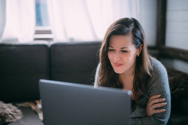 Eine Frau sitzt mit einem Laptop im Wohnzimmer, blickt auf den Bildschirm, macht sich Notizen, lernt eine Fremdsprache im Internet, liest einen Online-Kurs, unterrichtet sich im Internet, berät sich mit einem Kunden per Video - Foto, Bild