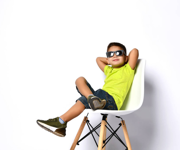 Kid in sunglasses, yellow t-shirt, denim shorts, khaki sneakers. Smiling showing victory sign, sitting on chair. Isolated on white - Photo, image