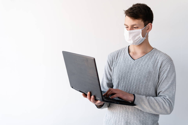 Nuori mies lääketieteellisessä naamiossa seisoo kotona kannettava tietokone kädessään. Suojaava lääketieteellinen naamio on pukeutunut hänen kasvoilleen. Portret kotona. Coronaviruksen hallintakonsepti. Etäkaranteeni covid-19
 - Valokuva, kuva