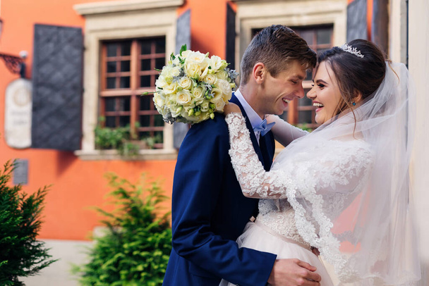 heureux jeunes mariés étreignant et souriant sincèrement. mariée tenant bouquet de mariage de roses. bâtiment avec fenêtres et volets en arrière-plan. - Photo, image