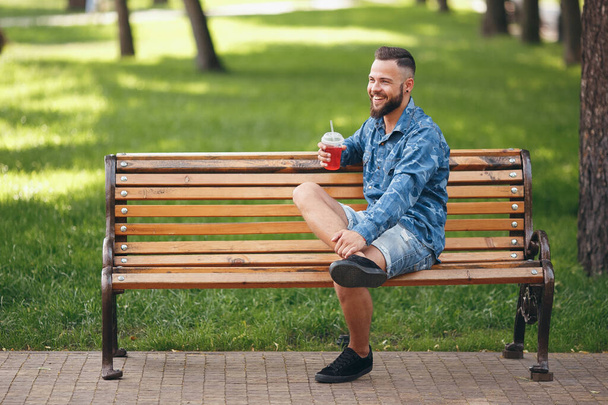 Een man met limonade rust in het voorjaar in een park op een bankje. Zonnige dag. Bladgroen. - Foto, afbeelding