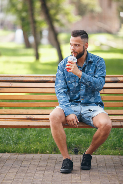 Mies limonadin kanssa lepää puistossa penkillä keväällä. Aurinkoinen päivä. Vihreät lehdet. - Valokuva, kuva