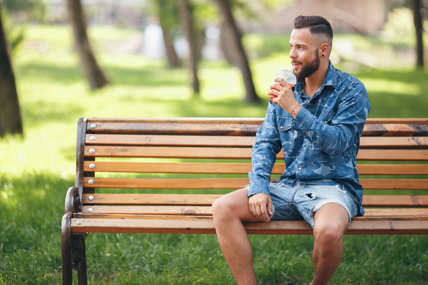 Ein Mann mit Limonade ruht sich im Frühling im Park auf einer Bank aus. Sonniger Tag. Grünes Laub. - Foto, Bild