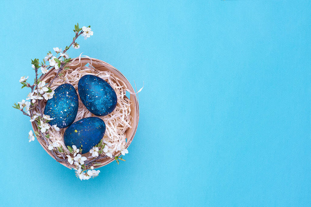 Huevos azules de Pascua en una canasta con flores sobre un fondo violeta. Fotografía horizontal. Vista superior Flatley. Vista superior. Objeto en el lado izquierdo de la foto
 - Foto, imagen