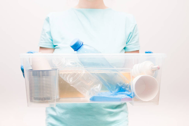 woman in a light blue t-shirt holds a plastic container with waste for recycling, waste-free life style, plastic bottles, a light bulb and a tin can, garbage sorting concept - Foto, afbeelding