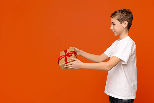 Netter Teenager-Junge gibt Geschenkbox beiseite, orange Hintergrund - Foto, Bild