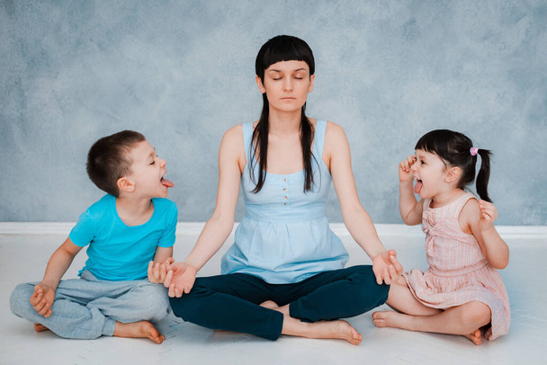 mom sitting floor grey blue background wall, meditating Lotus position, small children are screaming playing around focused calm mom meditating balancing - Photo, Image