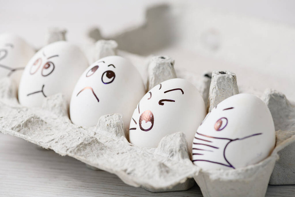 Huevos de pollo con caras de garabato con máscaras médicas en caja. Dibujado por la pluma marcador emociones en huevos blancos cáscara en máscaras para la epidemia 2020 coronavirus y vacaciones de Pascua en casa en aislamiento
. - Foto, Imagen