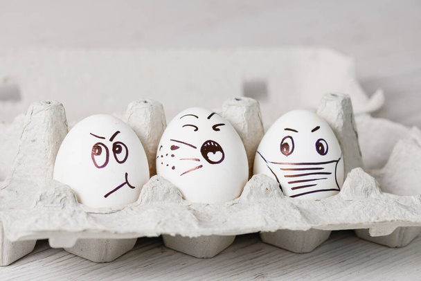 Painted by marker white eggs with funny emotional faces close up in paper tray for spring holiday during epidemic coronavirus. - Foto, Imagen