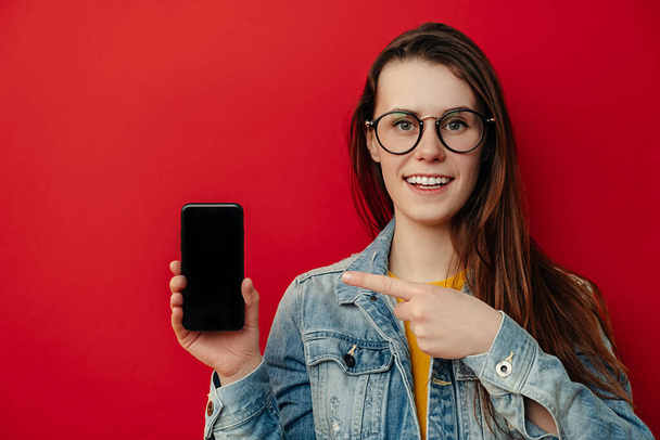 Smile young woman in spectacles, points at smart phone device, shows blank screen for your promotional content, wears denim jacket, advertizes new device for customers, isolated over red background - Foto, Bild