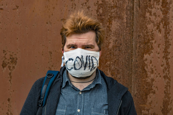 Kaukasische weiße Mann in der Hand gemacht rustikale Schutzmaske mit lebhaftem Text Blick auf Kamera auf rostige Wand depressive Hintergrundwand - Foto, Bild