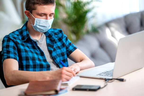 active teenager in a mask and headphones with homework on a laptop during a coronavirus quarantine. Social distance, self-isolation. Distance learning due to virus, COVID-19 - Zdjęcie, obraz