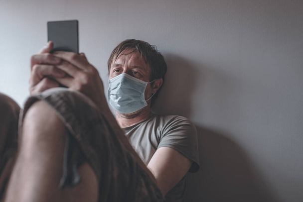 Man in self-isolation during virus outbreak using mobile phone. Worried male person with protective surgical mask, holding smartphone in stay at home concept, selective focus - Photo, image