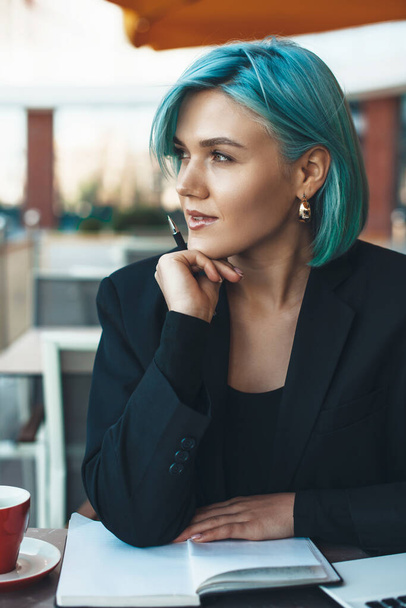 Empresaria de pelo azul mirando hacia otro lado mientras está sentada en una cafetería y leyendo un libro
 - Foto, imagen