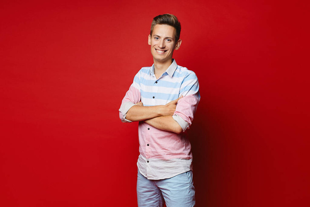 Handsome young man with modish hairstyle in shirt and shorts posing with folded hands on the red background - Φωτογραφία, εικόνα