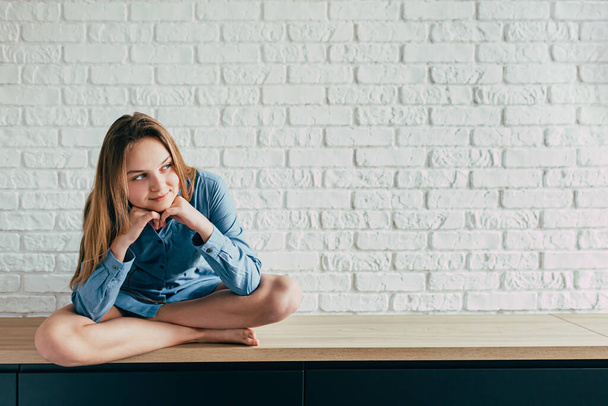 young girl in a blue shirt sits cross-legged against a large white brick wall and looks thoughtfully off to the side with a smile on her face. teenager with - Фото, изображение