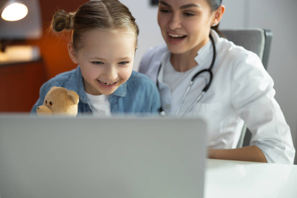 Grappig meisje glimlachen naar laptop in de buurt van kinderarts binnen - Foto, afbeelding
