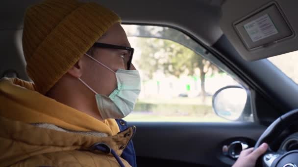 Handsome Caucasian man in winter yellow hat in a medical protective mask from virus on his face with black glasses drive a car and sneezes in hand on a bright sunny day close up slow motion - Footage, Video