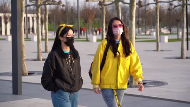 Ein paar süße Mädchen in schwarzen und gelben Jacken mit medizinischen Schutzmasken gehen abends an der frischen Luft auf die Straße. Frauen kümmern sich nicht um Quarantäne und Hausverbot. - Filmmaterial, Video