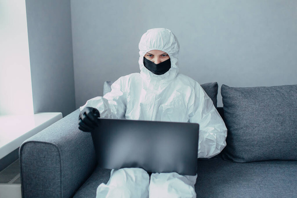 A nurse in a viral protective suit and black mask works on a laptop and looks for information about the virus epidemic. Quarantined because of the coronavirus epidemic.  - Фото, изображение