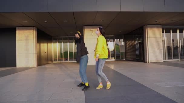 Ein paar glückliche attraktive Freundinnen in schwarzen und gelben Jacken tanzen fröhlich in medizinischen Schutzmasken vor dem Hintergrund des Gebäudes während der Quarantäne in etwa 19 Zeitlupe.. - Filmmaterial, Video