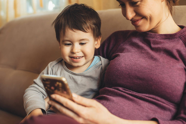 primer plano retrato de una madre caucásica embarazada pasar tiempo con su pequeño niño usando un teléfono en el sofá
 - Foto, Imagen