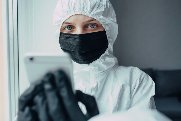 Doctor in viral protective suit wearing, medicine mask and gloves looks with fear at camera and hold phone quarantined worries about the spread of coronavirus.Virologist is afraid virus epidemic. - 写真・画像