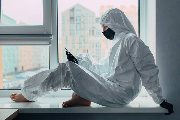 the girl is sitting on the window sill in face mask and viral protection suit, holding cellphone. Self quarantined due to coronavirus epidemic and isolated from the city. Social Distancing by Curfews - Photo, Image