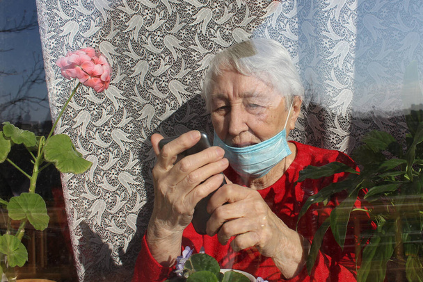 Ковід-19, охорона здоров'я, безпека та пандемія - стара самотня жінка, одягнена в захисну медичну маску, що сидить біля вікна будинку для захисту від вірусу за допомогою мобільного телефону. - Фото, зображення