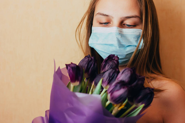 Joven adolescente caucásica en una máscara facial sostiene ramo de tulipanes en sus manos y esperar a la llegada de la primavera en cuarentena, ya que está auto-aislada debido al brote del coronavirus
. - Foto, imagen