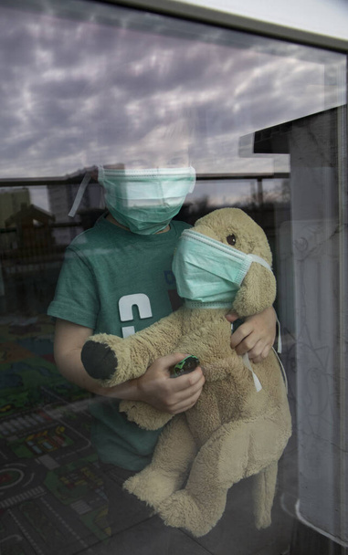 Coronavirus, Covid-19, Çocuk ve koruyucu maskeli oyuncak köpeği pencereden bakar, evdeki karantinaya bakar.  - Fotoğraf, Görsel